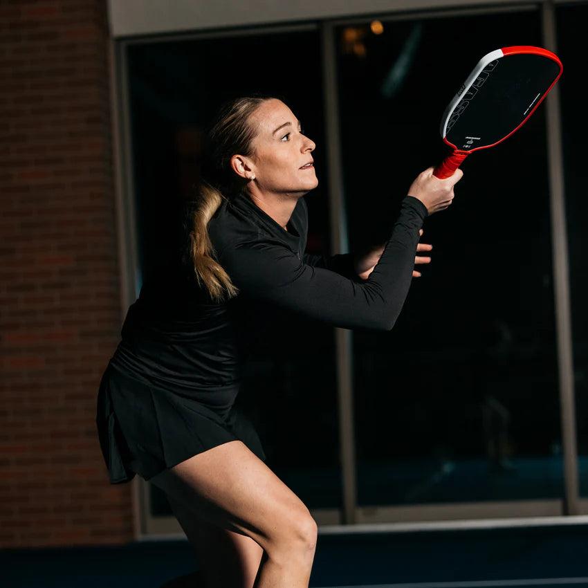 Woman in black sports clothing playing Pickleball with red bat in indoor setting.