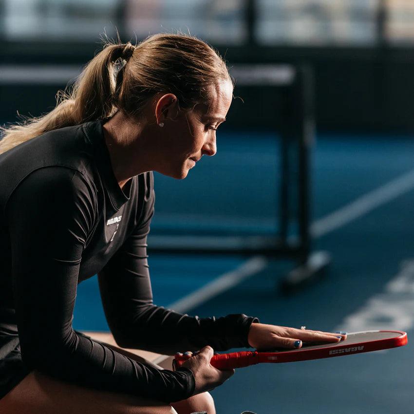Woman in sports clothing sitting with tennis bat in hand on a tennis court bench.
